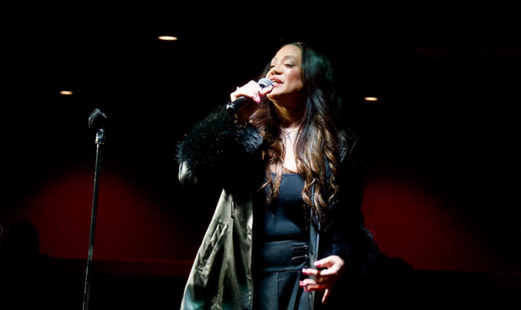 Medium-skin-toned woman sings into a microphone in a dark room.
