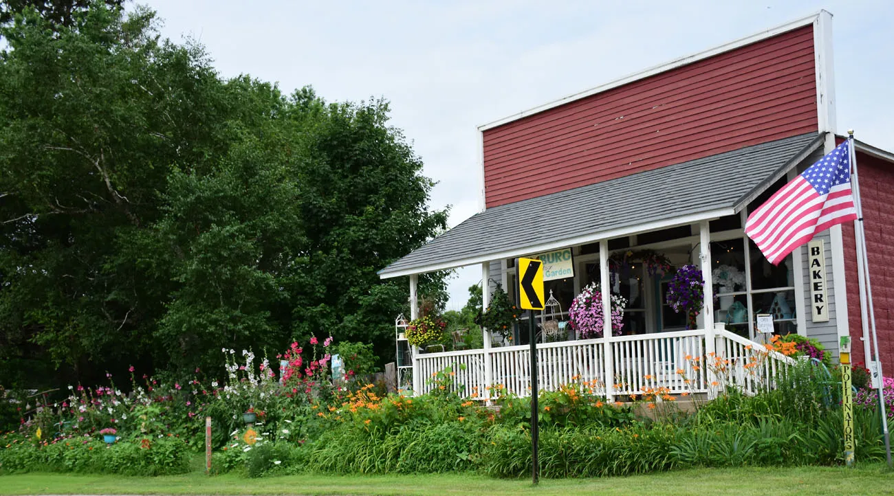 A red building with a white porch and shingled awning. There is a vertical sign on the right corner that says "Bakery." There is an American flag on a pole next to it. There is a garden filled with towering red and pink foxgloves, orange tiger lilies, and several hanging baskets filled with purple, pink, white, yellow and orange flowers.